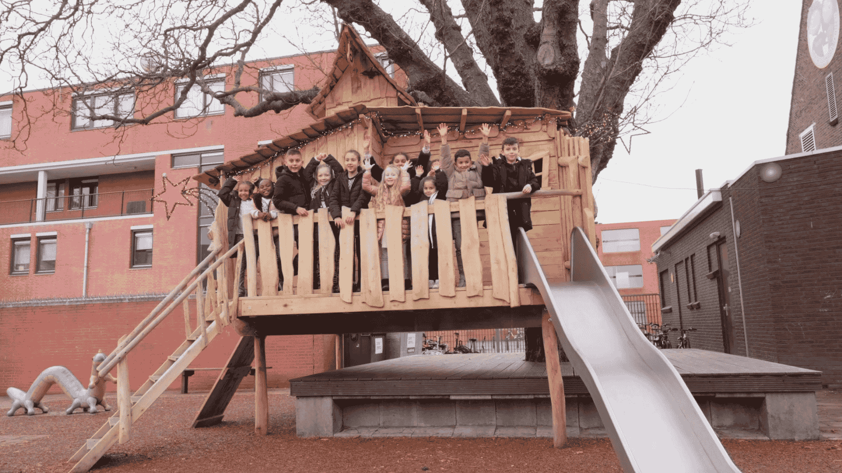 kinderen staan te zwaaien achter een balustrade van een boomhut. een grote boom met takken en een uitkijktoren is te zien op de achtergrond. Een glijbaan op de voorgrond en er is een valdempende ondergrond aangebracht onder de boomhut