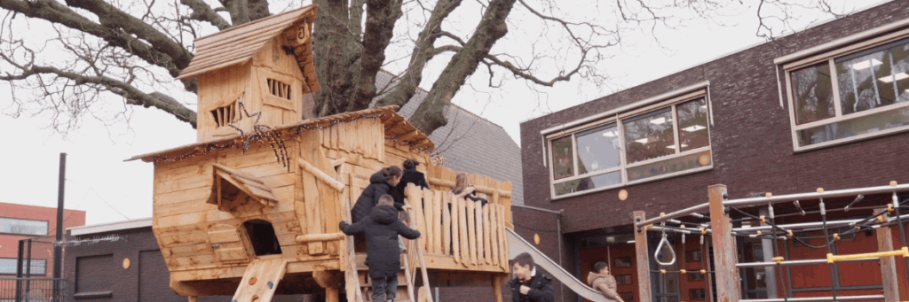 Openbaar speeltoestel boomhut, waar kinderen in aan het spelen zijn. Er is een boom op de achtergrond die geïntegreerd is in de boomhut op een schoolplein